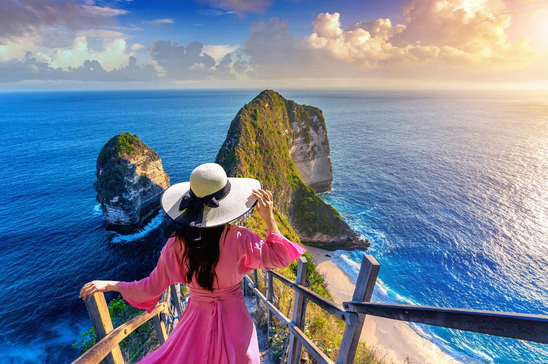 Woman in pink with a white hat overlooking the breathtaking Kelingking Beach on Nusa Penida Island, a must-visit during a Bali Tour
